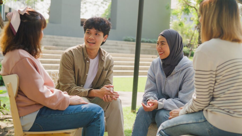 Four young adults in casual clothes, chatting outdoors in a park-like setting.